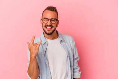 Pembe arka planda dövmeleri olan beyaz bir adam parmaklarıyla taş hareketi gösteriyor.