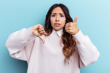 Mavi arka planda izole edilmiş genç Meksikalı kadın baş parmağını kaldırıyor, zor seçim konsepti.