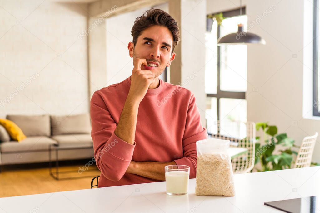 Young mixed race man eating oatmeal and milk for breakfast in his kitchen relaxed thinking about something looking at a copy space.