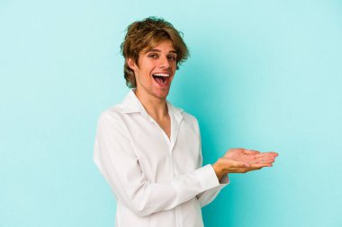 Mavi arka planda izole edilmiş beyaz makyajlı genç bir adam palmiye ağacında bir kopya alanı tutuyor..