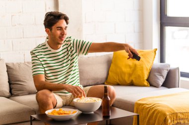 Genç melez adam kanepede oturmuş patlamış mısır yiyor.