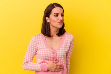 Sarı arka planda izole edilmiş genç beyaz kadın karnına dokunuyor, nazikçe gülümsüyor, yemek yiyor ve tatmin oluyor..