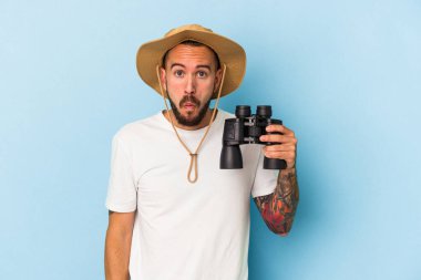 Elinde dürbün tutan beyaz bir genç adam mavi arka planda izole edilmiş omuz silkiyor ve gözleri açık..