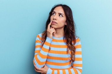 Mavi arka planda izole edilmiş genç Meksikalı kadın şüpheli ve şüpheci bir ifadeyle yana bakıyor..