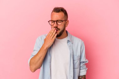 Pembe arka planda izole edilmiş beyaz dövmeli genç bir adam esniyor. Ağzı eliyle kapatan yorgun bir hareket sergiliyor..