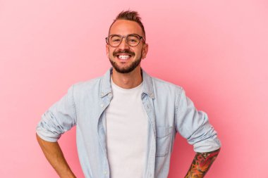 Pembe arka planda izole edilmiş beyaz dövmeli genç bir adam. Kendine güveniyor. Kalçalarını tutuyor..