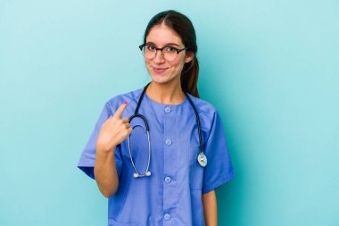 Kafkasyalı genç hemşire mavi arka planda izole edilmiş. Sanki seni davet ediyormuş gibi parmağıyla işaret ediyor..