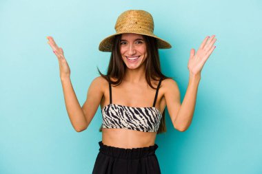 Genç, beyaz bir kadın kumsala gidiyor. Mavi arka planda izole edilmiş bir bikini giyiyor. Hoş bir sürpriz, heyecanlı ve el kaldırıyor..