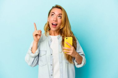 Elinde cep telefonu olan beyaz bir kadın mavi arka planda izole edilmiş ve ağzı açık bir şekilde üst tarafı işaret ediyor..