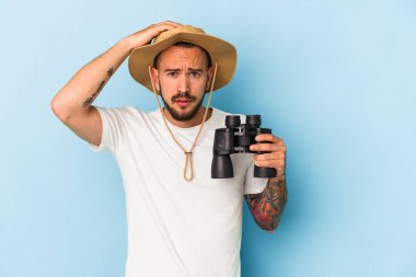 Mavi arka planda izole edilmiş dürbünleri olan beyaz dövmeli genç adam şoka uğruyor..