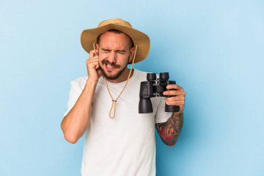 Dürbünlü dövmeleri olan genç beyaz bir adam mavi arka planda izole edilmiş kulakları elleriyle kapatıyor..