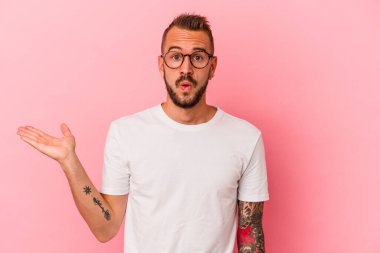 Pembe arka planda izole edilmiş dövmeleri olan beyaz genç bir adam, avuç içi fotokopi alanını elinde tutmaktan etkilenmiş..