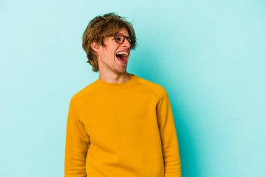 Makyajlı genç beyaz adam mavi arka planda izole edilmiş rahat ve mutlu bir şekilde gülüyor, boynunda dişlerini gösteriyor..