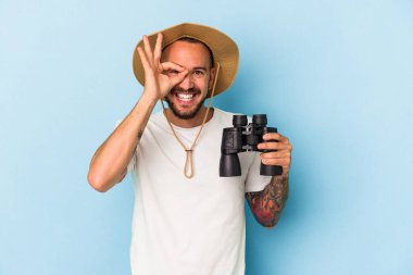 Mavi arka planda dürbün tutan beyaz dövmeli genç bir adam heyecanlı..