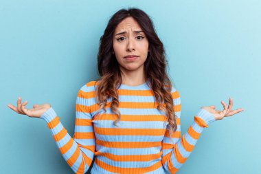 Meksikalı genç bir kadın mavi arka planda izole edilmiş. Sorgulama sırasında omuz silkiyor ve şüphe ediyor..
