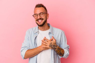 Pembe arka planda izole edilmiş dövmeleri olan genç beyaz bir adam. Gülüyor, elini kalbe koyuyor, mutluluk kavramını koruyor..