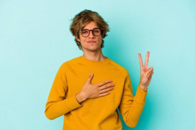 Makyajlı genç beyaz adam mavi arka planda izole edilmiş yemin ediyor, elini göğsüne koyuyor..