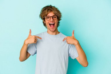 Mavi arka planda soyutlanmış beyaz makyajlı genç adam parmağıyla işaret ederek şaşırdı, geniş bir gülümsemeyle..