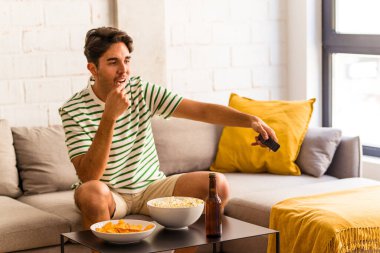 Genç melez adam kanepede oturmuş patlamış mısır yiyor.