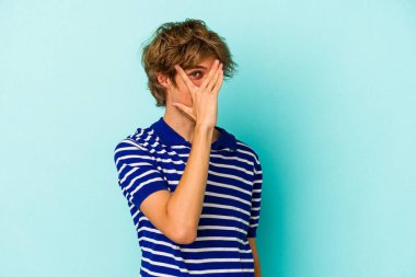 Makyajlı genç beyaz adam mavi arka planda izole edilmiş parmaklarıyla kameraya göz kırpıyor, yüzünü kapatmaktan utanıyor..
