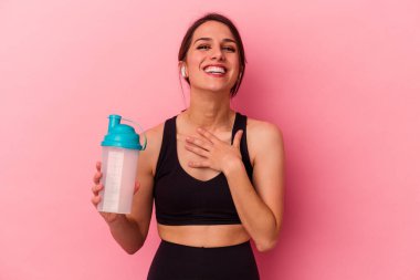 Pembe arka planda izole edilmiş protein içeceği içen beyaz bir kadın yüksek sesle gülüyor ve elini göğsüne koyuyor..