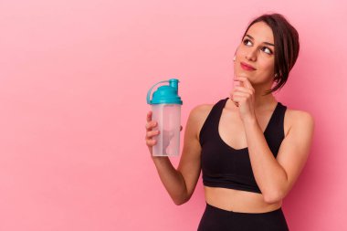 Pembe arka planda izole edilmiş protein içeceği içen beyaz bir kadın şüpheli ve şüpheci bir ifadeyle yana bakıyor..