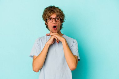 Makyajlı genç beyaz adam mavi arka planda izole edilmiş şans için dua ediyor, şaşırmış ve ağzını açıyor..