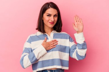 Pembe arka planda izole edilmiş genç beyaz kadın yemin ediyor, elini göğsüne koyuyor..