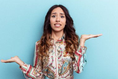 Mavi arka planda izole edilmiş genç Meksikalı kadın. Kopya alanını tutmak için kafası karışık ve omuz silkiyor..