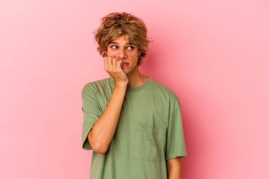 Makyajlı beyaz bir adam pembe arka planda izole edilmiş tırnaklarını yiyor, gergin ve endişeli..