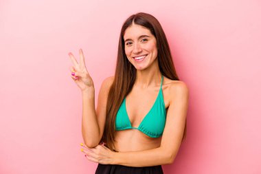 Bikini giyen beyaz bir kadın pembe arka planda izole edilmiş neşeli ve kaygısız parmaklarıyla barış sembolü gösteriyor..
