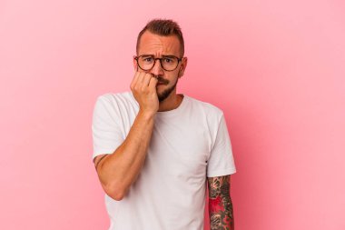 Pembe arka planda, tırnak yiyen, gergin ve endişeli dövmeleri olan genç beyaz bir adam..