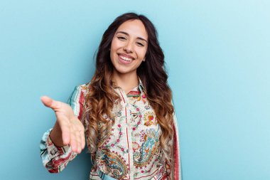 Mavi arka planda izole edilmiş genç Meksikalı kadın selamlama jesti olarak kameraya elini uzatıyor..