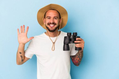 Mavi arka planda dürbün tutan beyaz dövmeli genç adam gülümseyerek beş numarayı parmaklarıyla gösteriyor..