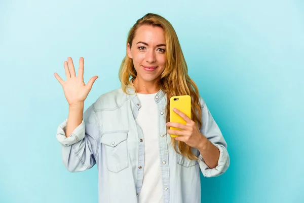 在蓝色背景下 拿着手机的高加索女人开心地微笑着 用手指拿着5号手机 — 图库照片