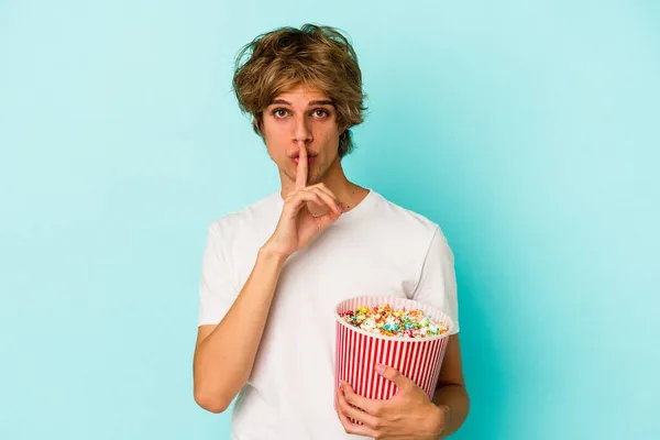 Young caucasian man with makeup holding popcorn isolated on blue background  keeping a secret or asking for silence.