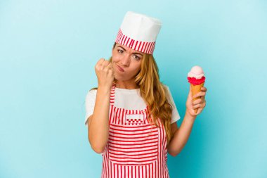 Kafkas dondurma üreticisi kadın elinde dondurma tutuyor mavi arka planda izole edilmiş yumruğunu kameraya gösteriyor, agresif yüz ifadesi var..