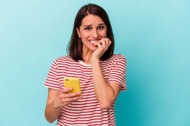 Elinde cep telefonu olan beyaz bir kadın mavi arka planda izole edilmiş tırnak yiyor, gergin ve endişeli..