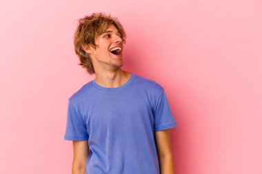 Makyajlı genç beyaz adam pembe arka planda izole edilmiş rahat ve mutlu bir şekilde gülüyor, boynunda dişlerini gösteriyor..