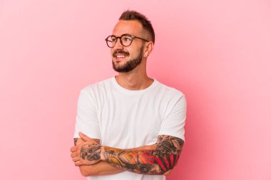 Pembe arka planda izole edilmiş dövmeleri olan genç beyaz adam kollarını kavuşturarak gülümsüyor..