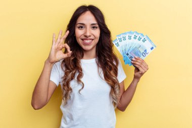 Sarı arka planda izole edilmiş faturaları tutan genç melez kadın neşeli ve kendinden emin, iyi bir jest yapıyor..