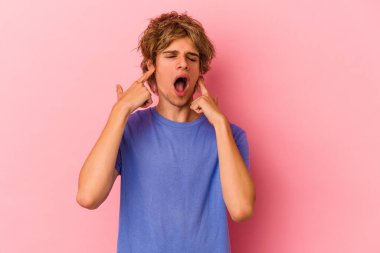 Makyajlı beyaz bir adam pembe arka planda izole edilmiş parmaklarıyla kulaklarını kapatıyor, gürültülü bir ortam yüzünden stresli ve çaresiz..