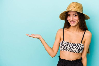 Mavi arka planda izole edilmiş bikini giyen beyaz bir kadın avucundaki fotokopi alanını ve beline bir el daha koyduğunu gösteriyor..