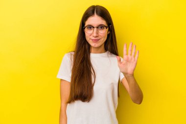 Sarı arka planda izole edilmiş genç beyaz kadın gülümseyerek beş numarayı parmaklarıyla gösteriyor..