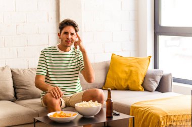 Genç melez adam kanepede oturmuş patlamış mısır yiyor. Parmağıyla şakağı işaret ediyor, düşünüyor ve bir göreve odaklanıyor..