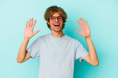 Makyajlı genç beyaz adam mavi arka planda izole edilmiş. 10 numarayı elleriyle gösteriyor..