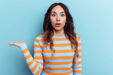 Mavi arka planda izole edilmiş genç Meksikalı kadın avuç içi fotokopi alanı tutmaktan etkilenmiş..