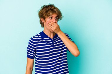 Makyajlı beyaz bir adam mavi arka planda izole edilmiş. Ağzını eliyle kapatan bir fotokopi odasına bakıyor..