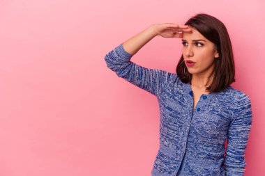 Pembe arka planda izole edilmiş genç beyaz kadın uzaklara bakıyor ve elini alnına koyuyor..