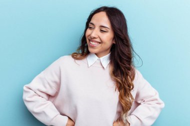 Mavi arka planda izole edilmiş genç Meksikalı kadın mutlu bir şekilde gülüyor ve ellerini karnının üstünde tutarak eğleniyor..